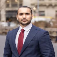 Professional man in a navy suit and red tie posing outdoors in an urban street setting.